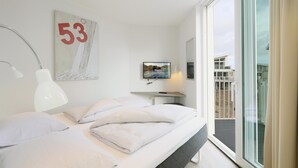 A modern bedroom with a bed, a desk, a TV, and a large window with a view of a building.