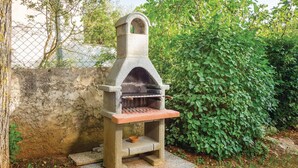 A stone-built outdoor grill with a simple design, situated in a garden with dense foliage and a chain-link fence in the background.