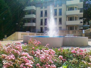 A fountain surrounded by pink flowers in front of a multi-story building with balconies.