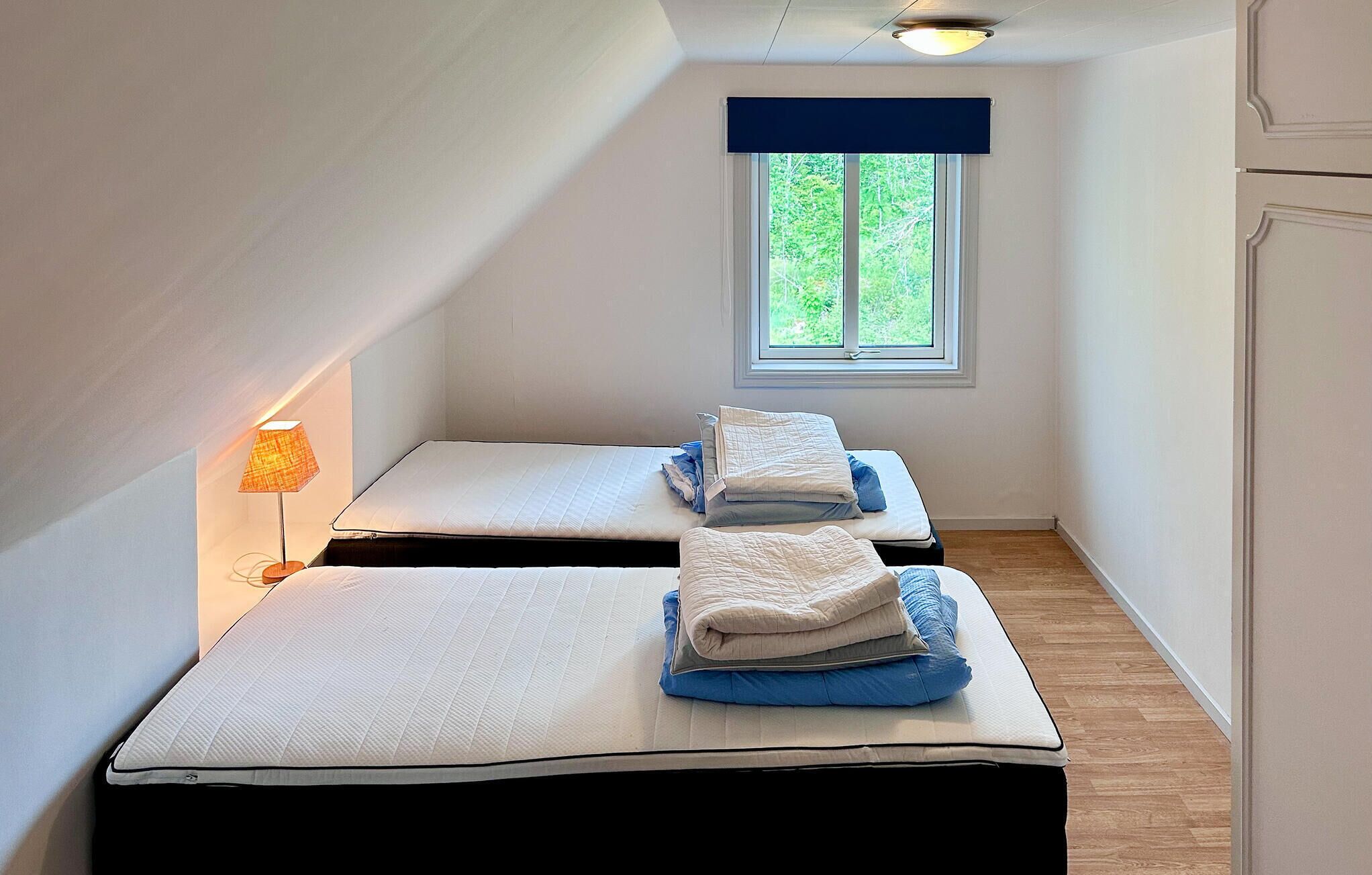 A small, narrow room with two beds, a window with a view of greenery, and a lamp on the left.