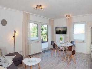 Ein modernes Wohnzimmer mit einer Couch, einem Esstisch mit Stühlen, einem an der Wand montierten Fernseher und großen Fenstern.