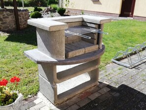 A concrete outdoor grill with a metal rack, set on a brick patio with a stone wall and grass in the background. - Vacation apartment Leentje on the ground floor - Ferienwohnungen Wiesenblick (Trassenheide)