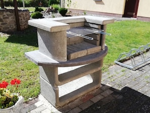 Un barbecue extérieur en béton, doté d’une grille en métal, installé sur une terrasse en briques, avec un mur en pierre et de l’herbe en arrière-plan.