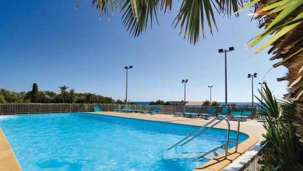 Une piscine d’un bleu éclatant, dotée d’une terrasse ensoleillée, entourée de palmiers et offrant une vue sur l’océan.