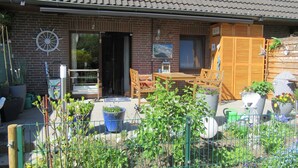 Ein Backsteinhaus mit Ziegeldach, einer Holztür und einer kleinen Terrasse mit Tisch und Stühlen. Kübelpflanzen und ein umzäunter Gartenbereich sind ebenfalls vorhanden.