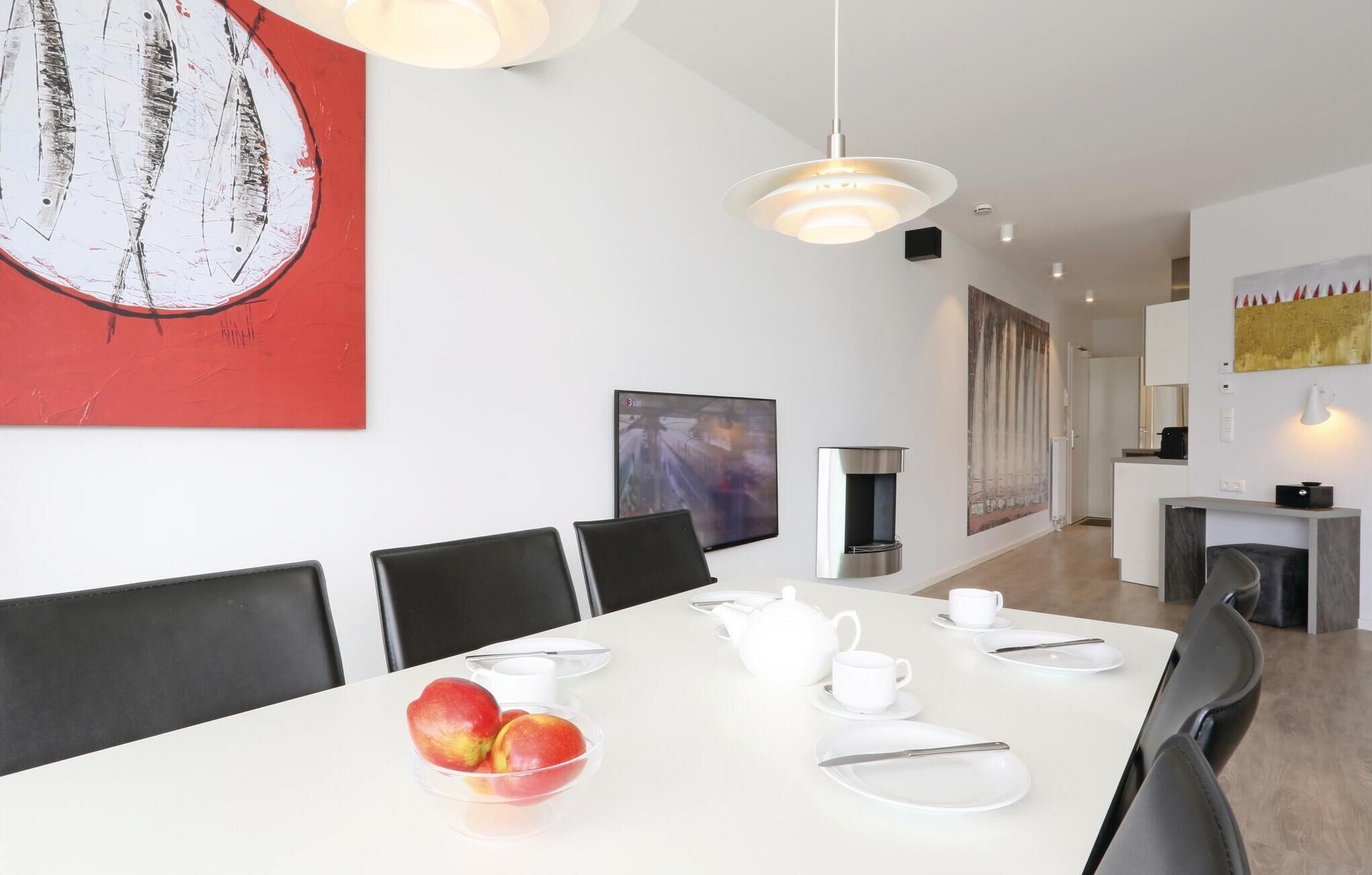 Een moderne eetruimte met een witte tafel voor vier personen, een schaal met perziken en een koffieapparaat.