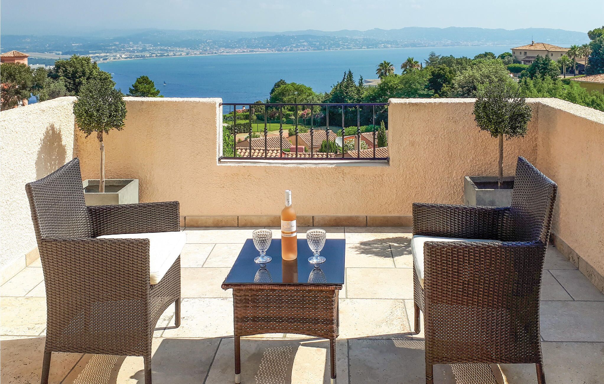 A terrace with wicker furniture, a table, and a bottle of wine, overlooking a coastal cityscape.