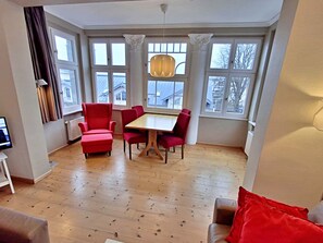 Una habitación con piso de madera, una mesa de comedor con sillas, un televisor y ventanas amplias con cortinas.