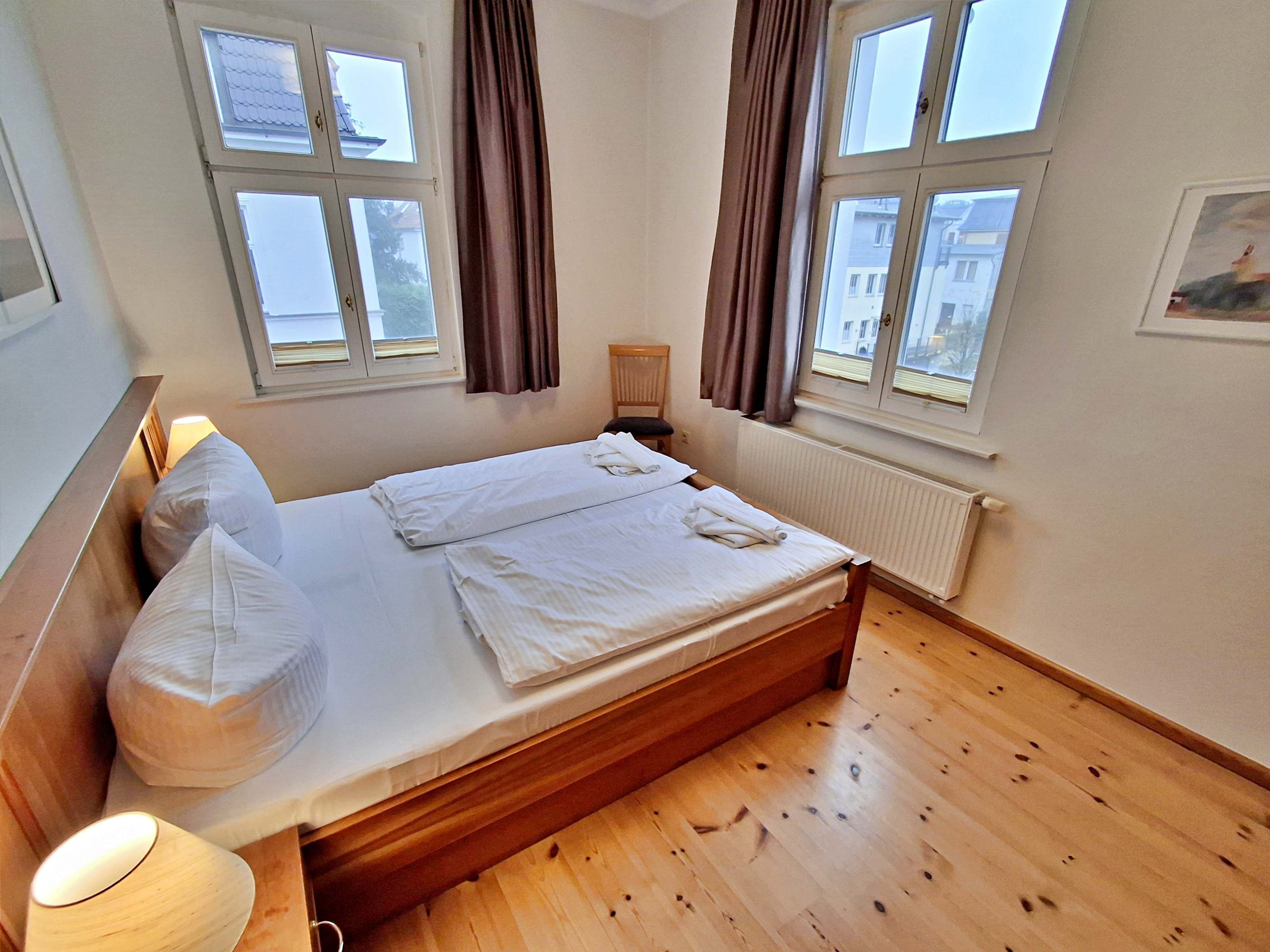 A bedroom with a wooden bed, two windows with curtains, a radiator, and a lamp.