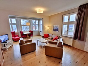 A spacious living room with a large window, a dining area with red chairs, a sofa, and a television. - Prerow - Villa Arlberghaus 1 (Heringsdorf)
