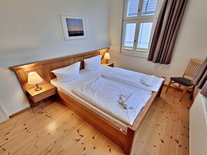 Una cama bien hecha con sábanas blancas, una lámpara de noche, una mesita de madera y una silla.