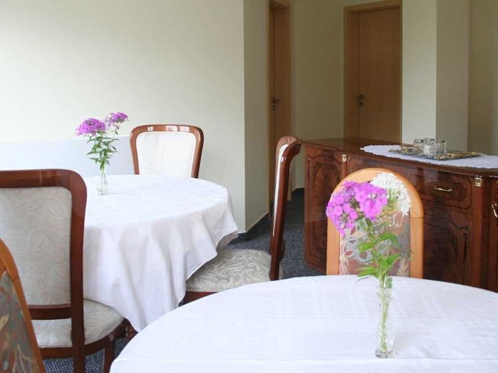 Een eetgedeelte met twee ronde tafels, elk met een witte tafelkleed en een vaas paarse bloemen. De tafels worden omringd door houten stoelen met beklede zitplaatsen.