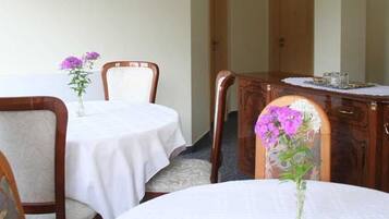 Een eetgedeelte met twee ronde tafels, elk met een witte tafelkleed en een vaas paarse bloemen. De tafels worden omringd door houten stoelen met beklede zitplaatsen.