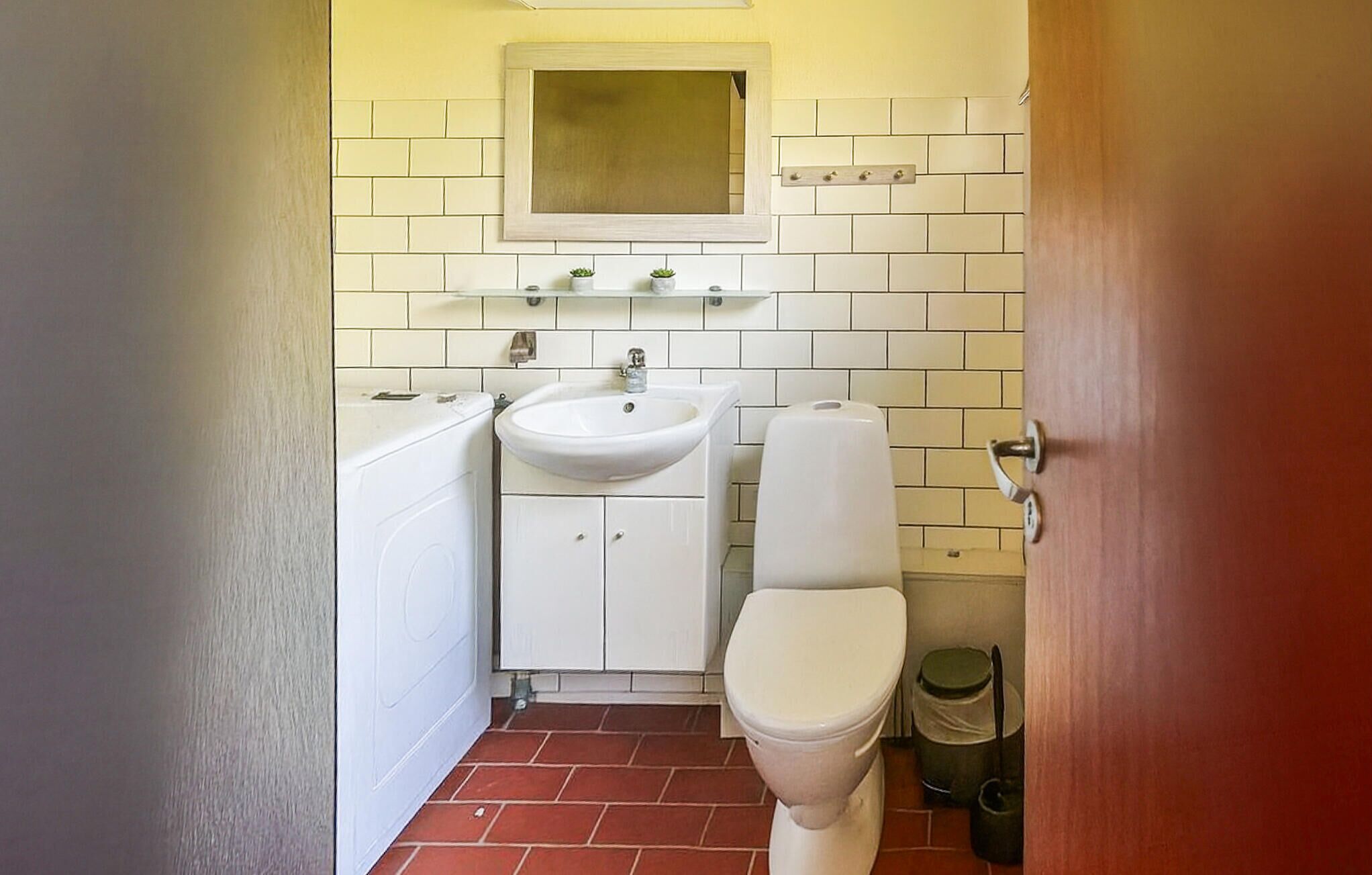 A small bathroom with a white sink, toilet, and a mirror above the sink. There are two small potted plants on the shelf above the sink.