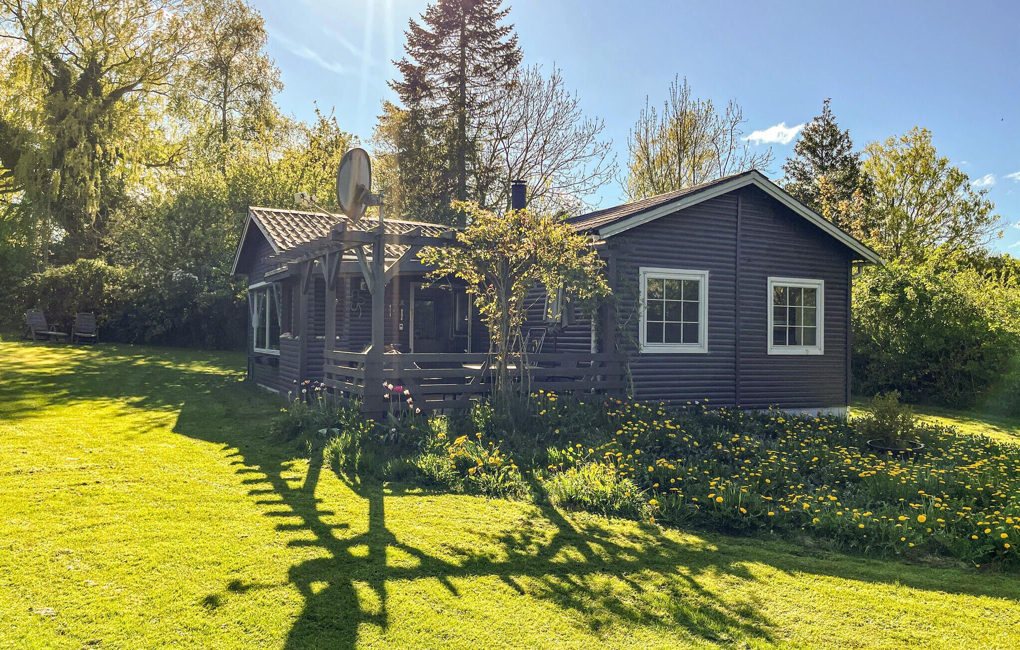 A log cabin with a porch and a satellite dish, surrounded by a well-maintained lawn and flowering plants.