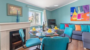 A dining area with a round glass table, blue chairs, and a colorful table runner. A fireplace is visible on the left, and a sofa with colorful cushions is on the right.