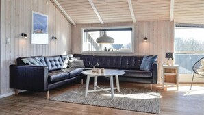 A modern living room with a dark blue sectional sofa, a round coffee table, and a large window with a view of the outside. - Nice home in Hvide Sande with sauna (Hvide Sande)