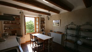 A kitchen with wooden beams, a dining table, chairs, shelves with dishes, and a window with curtains. - Tour Ourne Mas de Tours (Tornac)