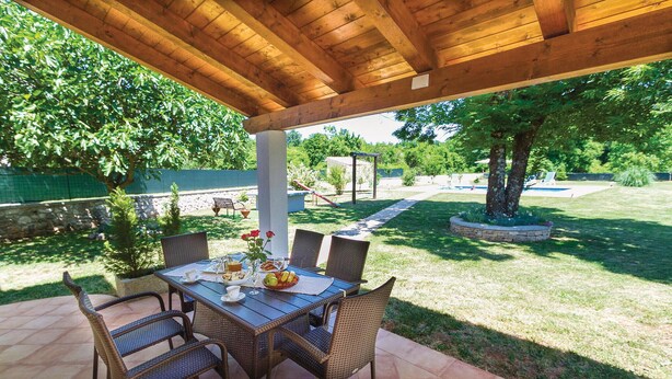 Una terrazza esterna con un pergolato in legno, un tavolo apparecchiato per mangiare, sedie in vimini e vista sul giardino.