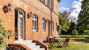A brick building with a stone staircase leading to a door, a table with chairs, and a garden in the background.