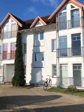 Um edifício residencial de vários andares com entrada central, uma bicicleta estacionada na frente e uma área de jardim bem cuidada.