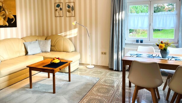 A living room with a sofa, a coffee table with fruit, a floor lamp, a dining table with chairs, and a window with curtains. - Fewo ivy - House Gundelrebe (Bergen auf Rügen)