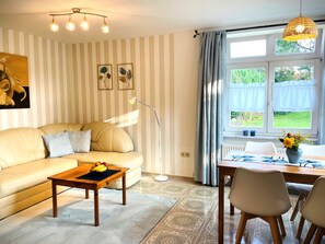 Un salon comprenant un canapé, une table basse avec des fruits, une lampe sur pied, une table à manger avec des chaises et une fenêtre avec des rideaux.