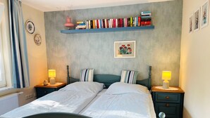A bedroom with a bed, two bedside tables, a bookshelf with books, a framed picture, and a radiator. - Fewo ivy - House Gundelrebe (Bergen auf Rügen)
