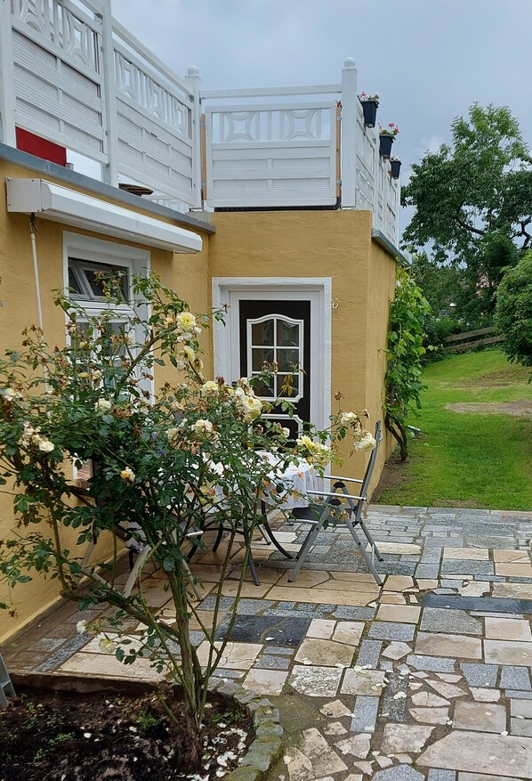 A yellow building with a white door, a balcony with potted plants, and a stone patio with a chair.