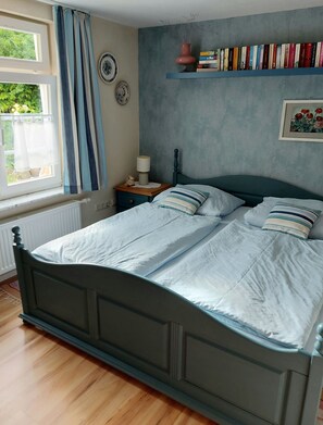 A bedroom with a bed, a nightstand, a window with curtains, a radiator, a wall shelf with books, and a framed picture.