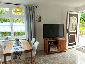 A room with a dining table, chairs, a television on a wooden stand, a door with a glass panel, and a window with curtains.