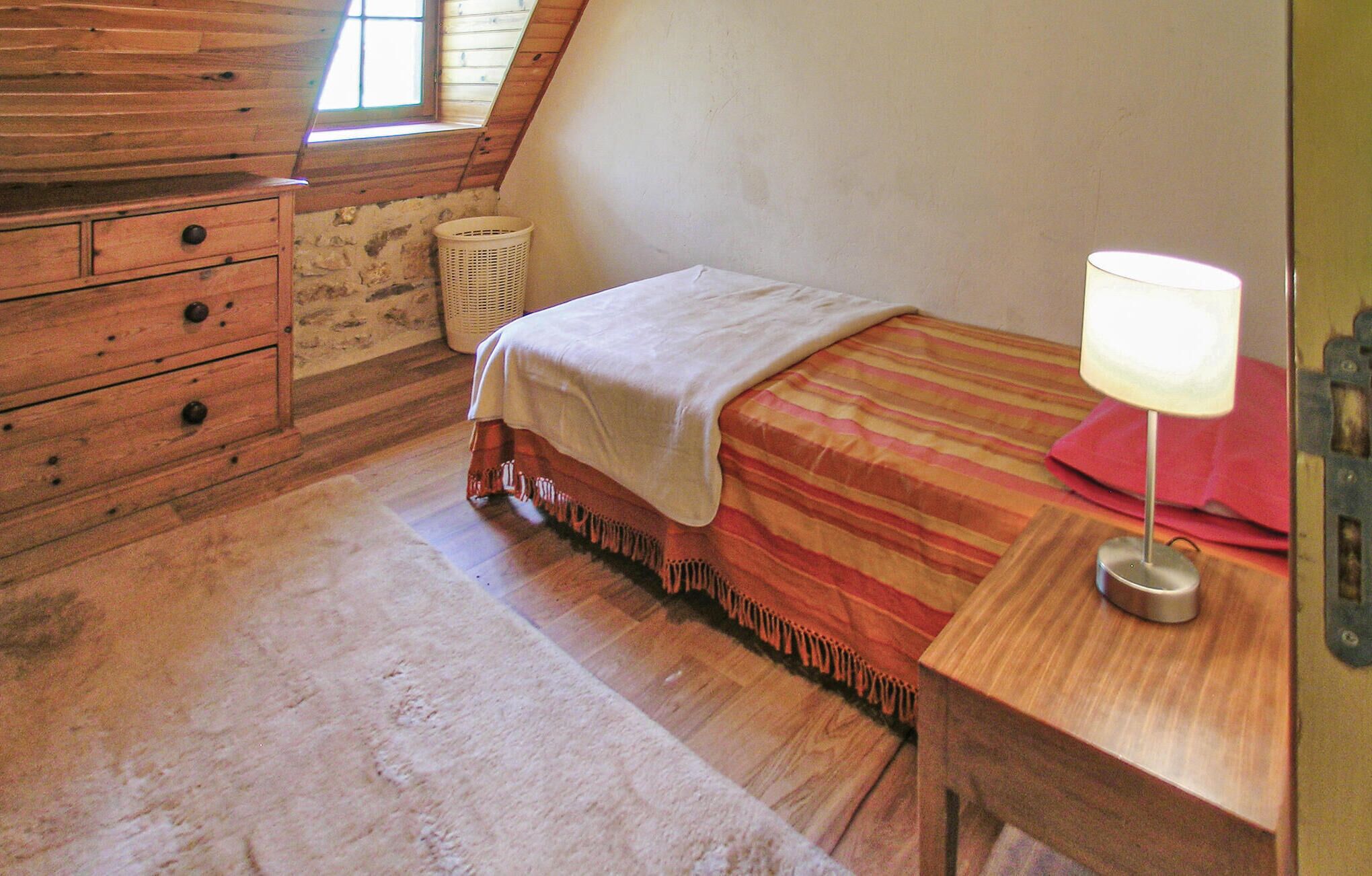 A bedroom with a wooden dresser, a bed with a striped bedspread, a bedside lamp, and a window with a view of the outside.