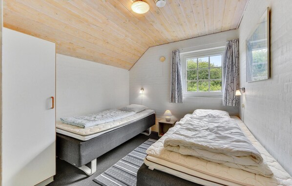 Une petite chambre à coucher accueillante, avec un plafond en pente, un lit simple, une table de chevet avec une lampe et une fenêtre avec des rideaux.