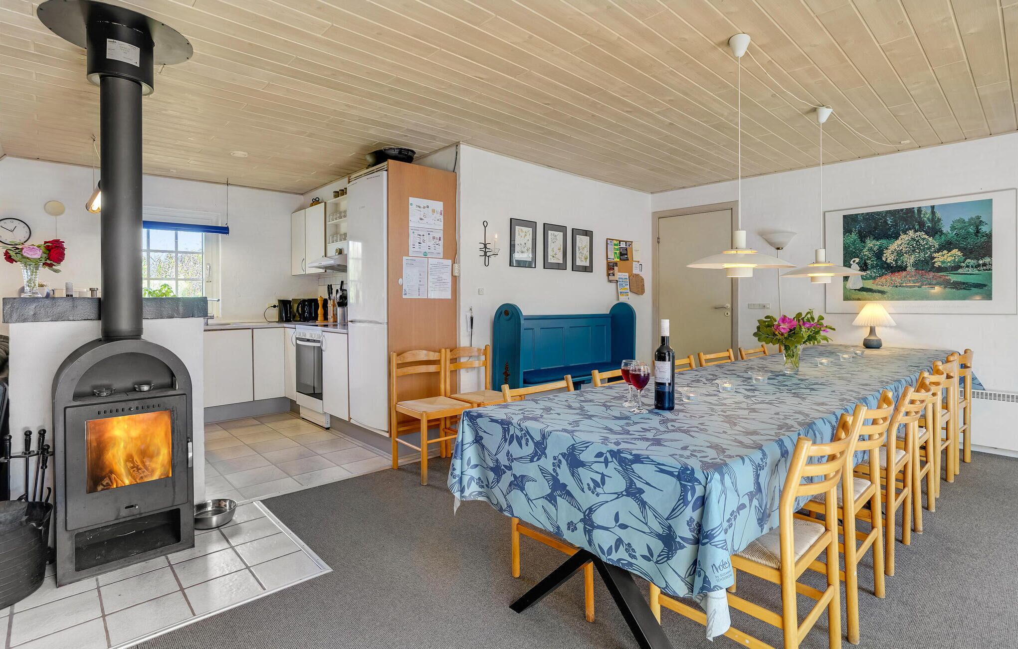 A cozy kitchen with a wood-burning stove, a dining area with a table set for six, and a blue patterned tablecloth.