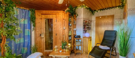 Un intérieur en bois avec un ventilateur de plafond, une porte en bois, une petite table avec des verres à vin, un fauteuil noir et des plantes en pot.