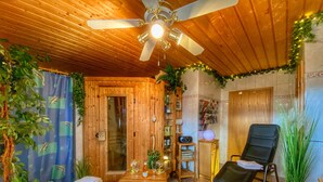 A wooden interior with a ceiling fan, a wooden door, a small table with wine glasses, a black lounge chair, and potted plants.