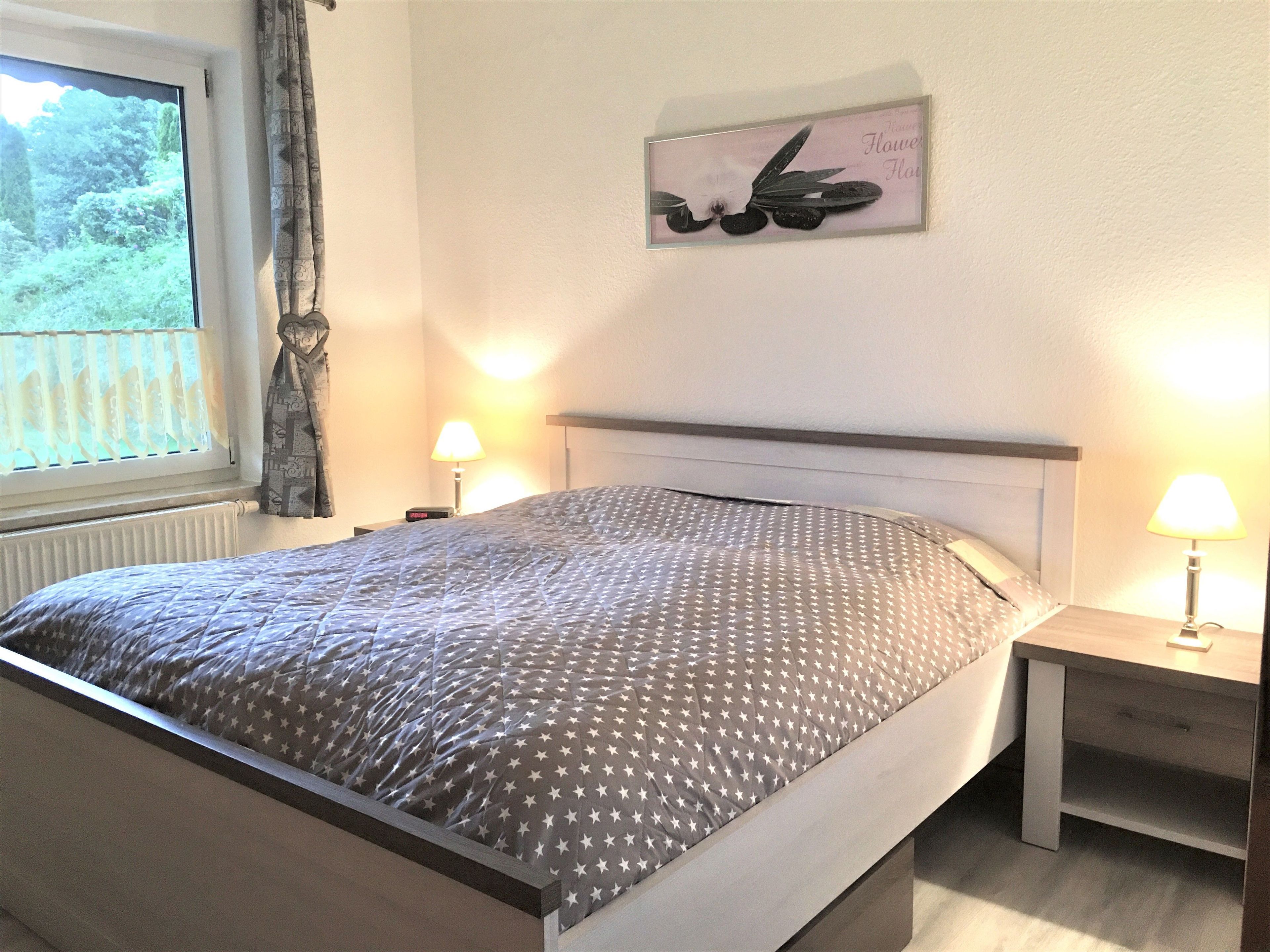 Un dormitorio con cama, mesitas de noche, ventana con cortinas e imagen colgada en la pared.