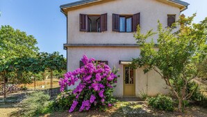 A two-story house with a yellow door, brown shutters, and a garden with purple flowers.
