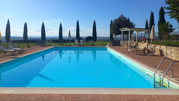 Une piscine rectangulaire à l’eau d’un bleu limpide, entourée d’une terrasse en briques. On y trouve des chaises longues et un espace salon couvert avec des parasols. De grands cyprès bordent l’arrière-plan, et un mur de pierre délimite l’espace piscine.