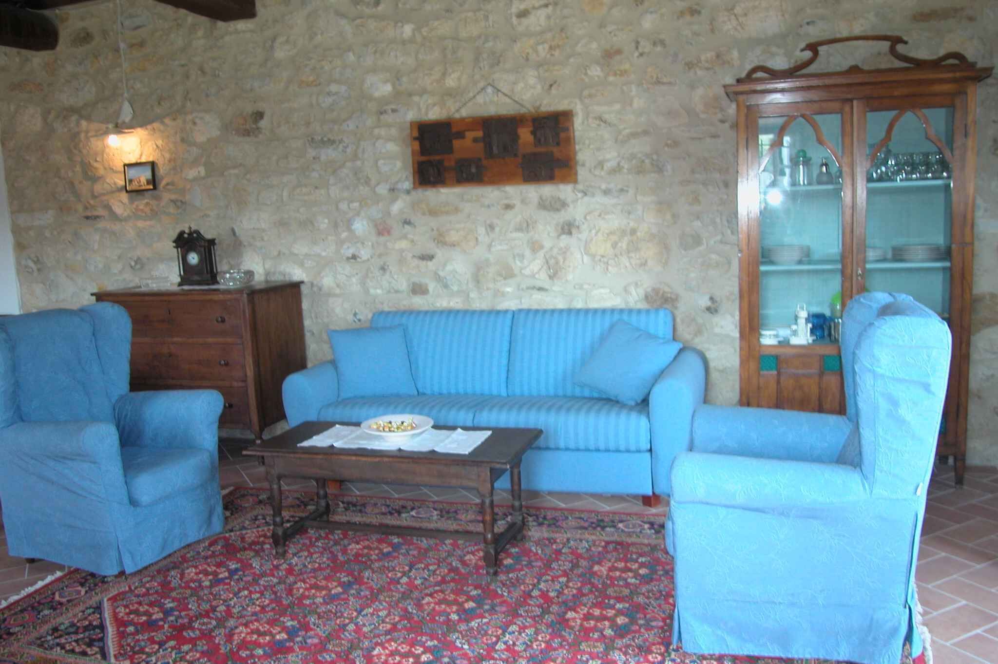 A living room with a stone wall, a blue sofa, two blue armchairs, a wooden cabinet, and a patterned rug.