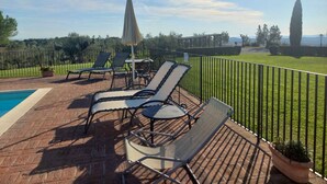 Un espace aménagé au bord de la piscine, avec des chaises longues, une table et une vue sur un champ verdoyant et des arbres.