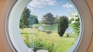 Vista a través de una ventana de un jardín con un estanque, setos recortados y diversas especies de árboles.