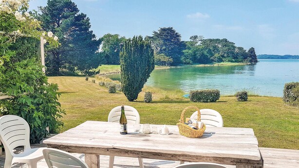 Terraza de madera con mesa y sillas con vista a un lago, rodeada de follaje y árboles.
