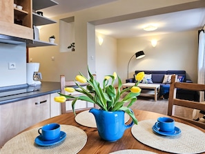 A modern kitchen with a coffee machine, a dining table with blue cups and a blue pitcher, and a living area with a sofa and a lamp.