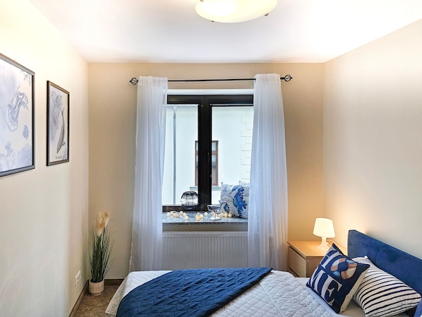 A bedroom with a bed, a window with curtains, a bedside table with a lamp, and framed artwork on the wall.