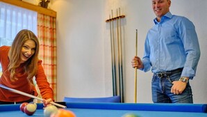 Une table de billard avec des billes et des queues, une fenêtre avec des rideaux et un porte-objets fixé au mur.
