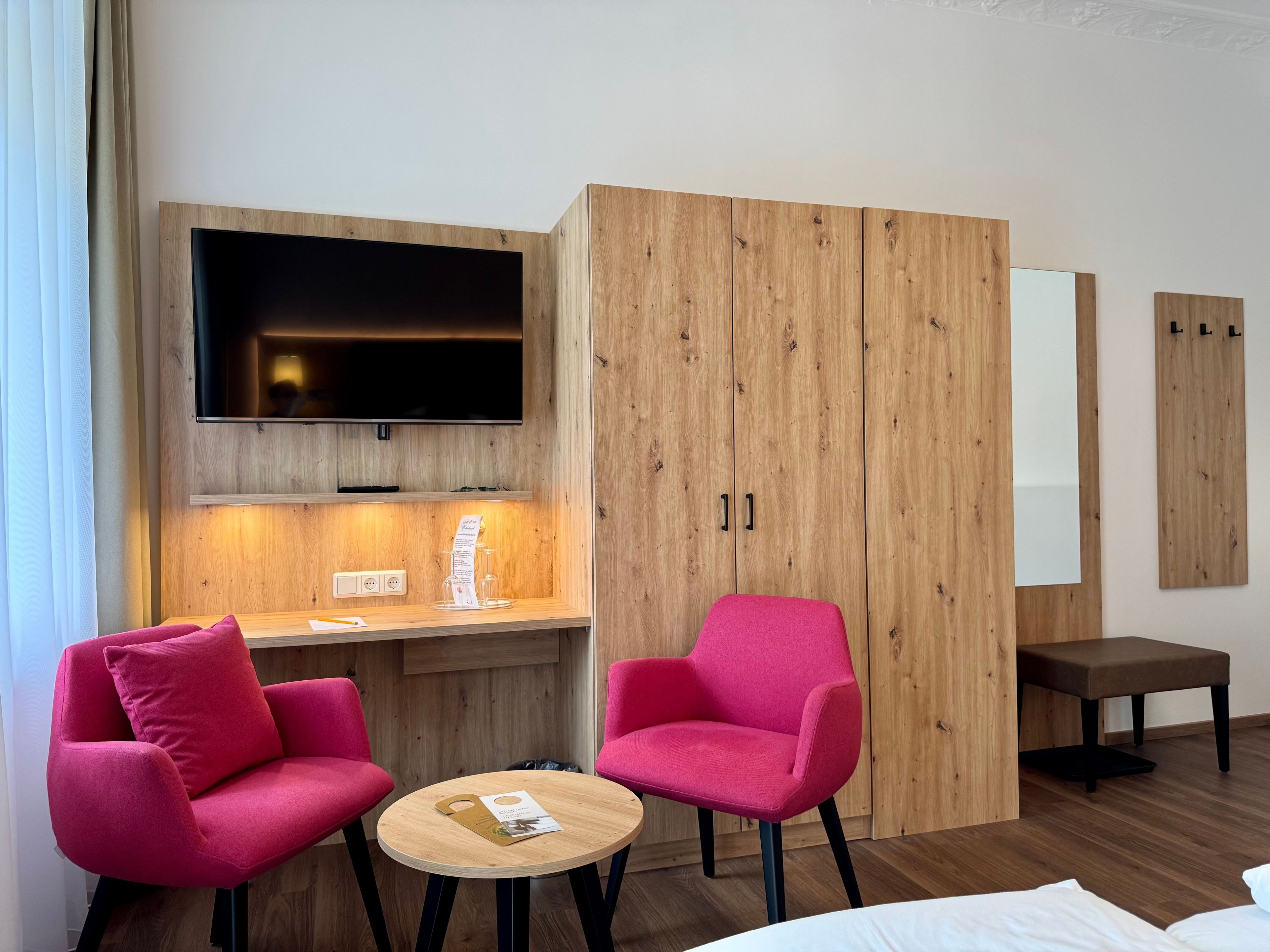 A hotel room with a wooden wardrobe, a flat-screen TV, a desk with a menu, two pink chairs, a small round table, and a bed.