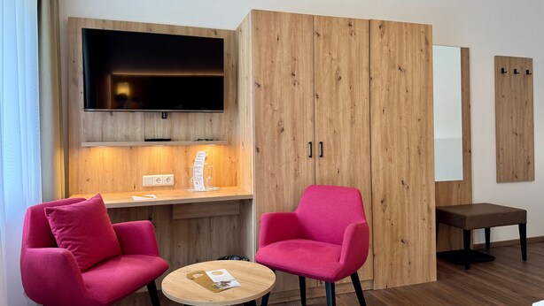 Une chambre d’hôtel avec une armoire en bois, un téléviseur à écran plat, un bureau avec un menu, deux chaises roses, une petite table ronde et un lit.