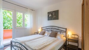 A double bed with white linens, two bedside tables with lamps, a window with sheer curtains, and a wall-mounted artwork. - Cozy home in Risika with WiFi (Risika)