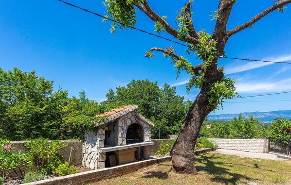 A stone-built outdoor kitchen with a stone chimney, surrounded by lush greenery and a large tree. - Cozy home in Risika with WiFi (Risika)